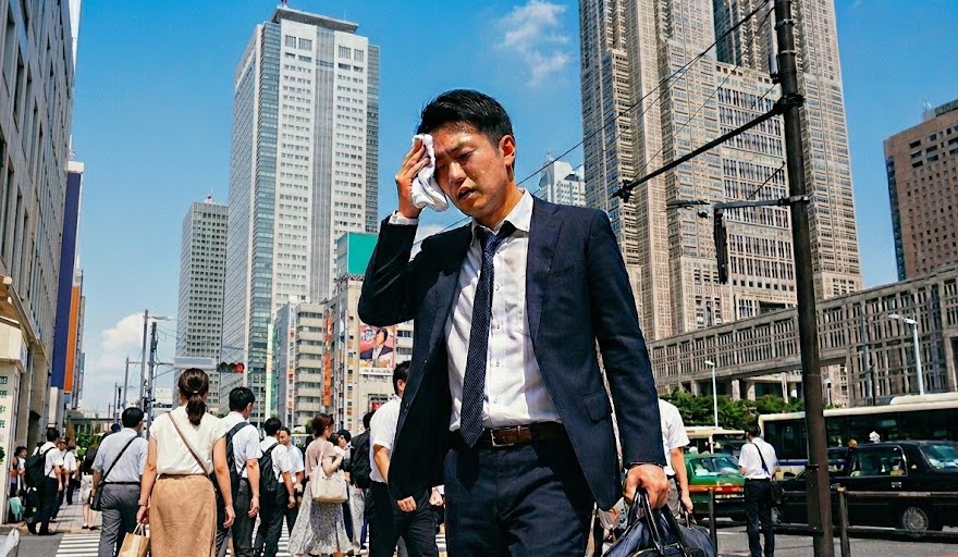 猛暑の東京・西新宿をスーツ姿で汗を拭いながら歩く28歳前後の転職活動中の男性。背景には青空と東京都庁や高層ビル群が広がる、夏の転職証明写真の服装マナーに関する記事のサムネイル画像。